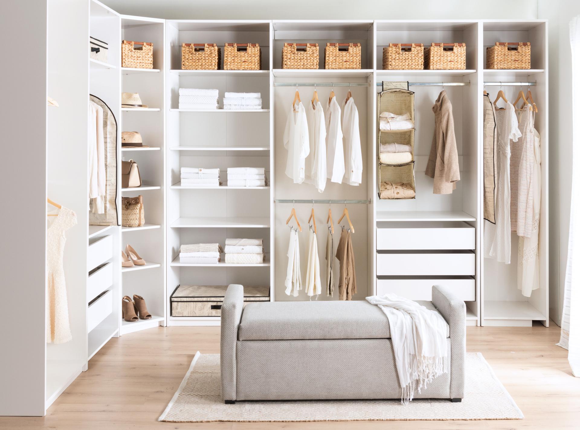 Organized walk-in closet system photograph featuring modular white shelving, hanging clothes, storage baskets, and a central gray ottoman bench.
